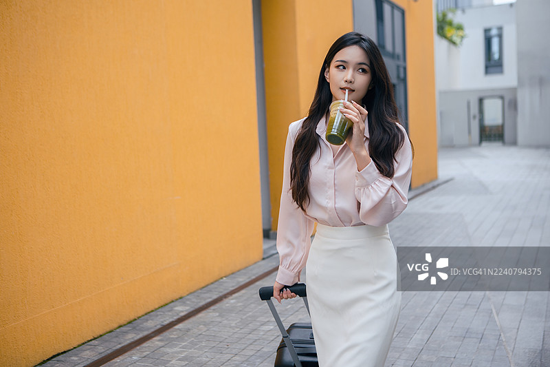亚洲女商人，手持果汁和行李箱，在有黄色墙壁的城市街道上旅行。职业女性企业家探索城市环境，将工作与休闲相结合，代表着现代商务旅行、独立和活力。图片素材