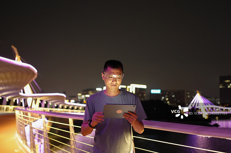 夜间，一名男子在河边戴着智能眼镜使用平板电脑。图片素材