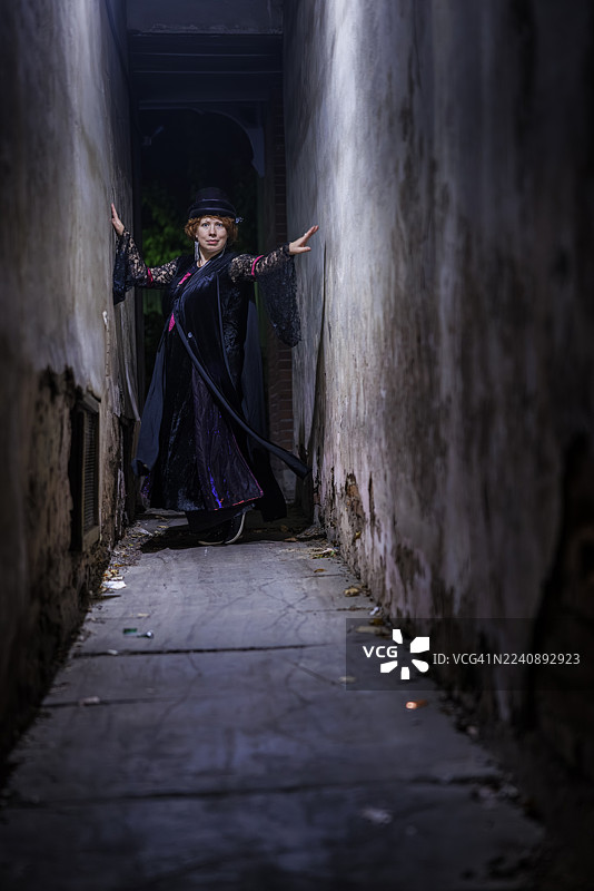 万圣夜，女巫穿行于黑暗狭窄的小巷。身穿维多利亚时期服装的红发女子在狭窄的城市通道中显露出担忧的神情。图片素材