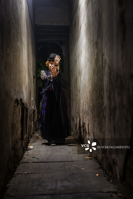神秘的红发女巫在黑暗的小巷中做出防御姿势。狭窄的城市通道中的万圣节人物。图片素材