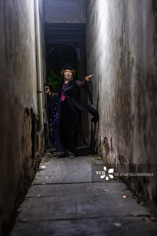 诡异的小巷里，一位中年女子身着女巫服装，正在戏剧化地挥舞手臂。万圣节角色从黑暗狭窄的通道中现身。图片素材