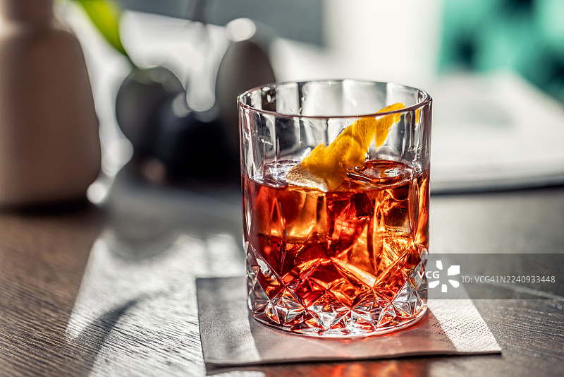 冰镇酒杯中盛着一杯优雅的内格罗尼鸡尾酒，并用橙子皮装饰。酒杯放在木桌的灰色餐巾上，一道明亮的 and 聚光灯戏剧性地照亮了酒，营造出温馨的氛围。图片素材
