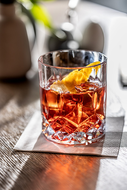 一款优雅的内格罗尼鸡尾酒盛放在冰霜感的玻璃杯中，并用橙皮装饰。杯子放在木桌的灰色餐巾上，一道明亮的 and dramatic light beam 照亮了酒，营造出温馨的氛围。图片素材