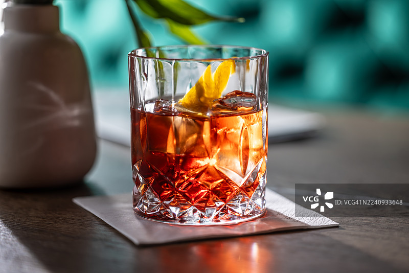 一款优雅的内格罗尼鸡尾酒盛放在冰镇酒杯中，并用橙皮装饰。酒杯放在木桌上的灰色餐巾上，一道明亮的 and 聚光灯光束戏剧性地照亮了酒，营造出温暖的氛围。图片素材