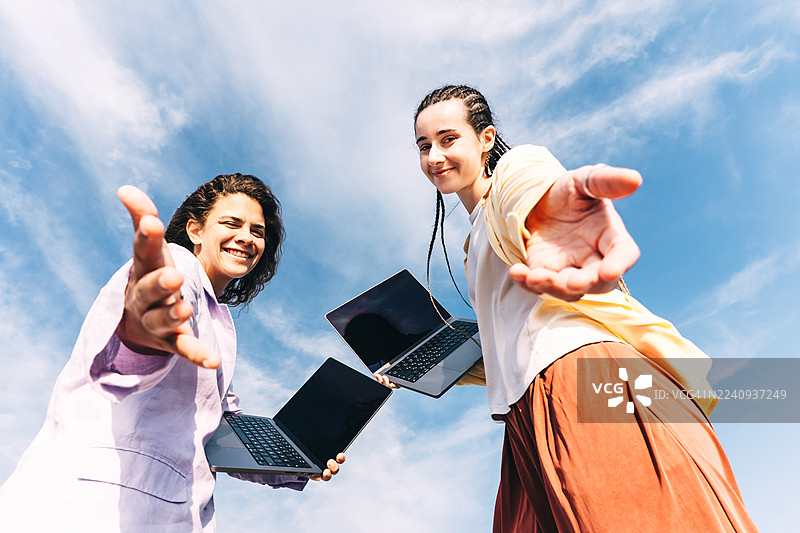 两位年轻女子在生机勃勃的蓝天下户外工作，她们面带微笑，向前伸出手。图片素材