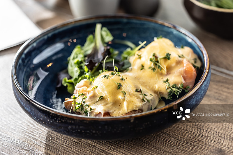 蓝色亮面陶瓷碗盛着的班尼迪克蛋。这道菜由两片面包组成，上面铺着烟熏三文鱼或火腿和水波蛋，淋上浓郁的黄色荷兰酱，并撒上新鲜的豆芽和香草。图片素材