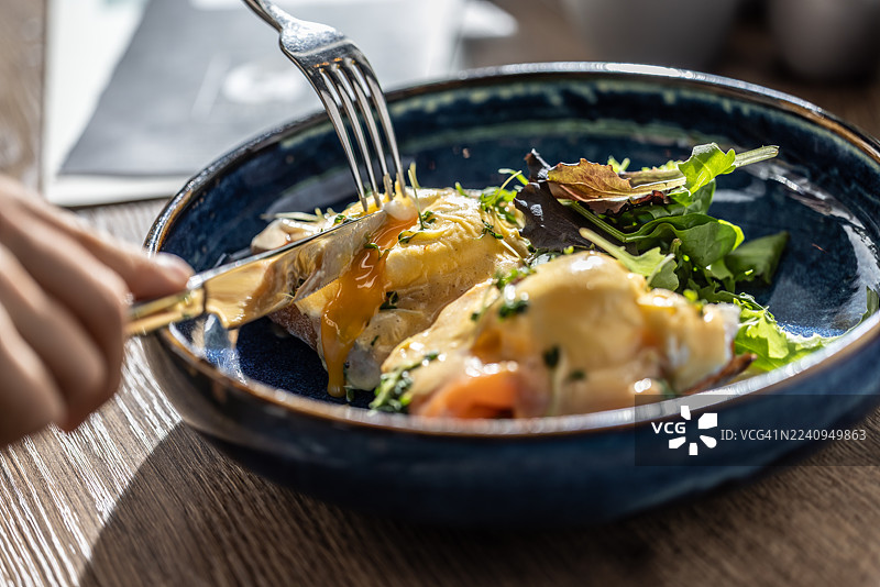 蓝釉陶瓷碗中的班尼迪克蛋，淋上荷兰酱，并用绿色香草点缀。餐叉和餐刀插入菜肴中，蛋黄流淌出明亮的黄色。图片素材