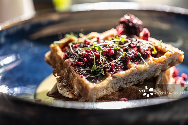 一个光亮的蓝色碗里盛着华夫饼，华夫饼分两层，上面淋着浅色奶油或酱汁，并用深红色撒料（可能是覆盆子或黑莓）和新鲜香草或嫩芽装饰。图片素材