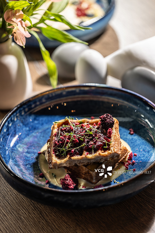 亮蓝色的闪亮碗里盛着华夫饼。两层华夫饼上面淋着浅色奶油或酱汁，并用深红色糖粒（可能是覆盆子或黑莓）和新鲜香草或豆芽装饰，十分丰富。图片素材
