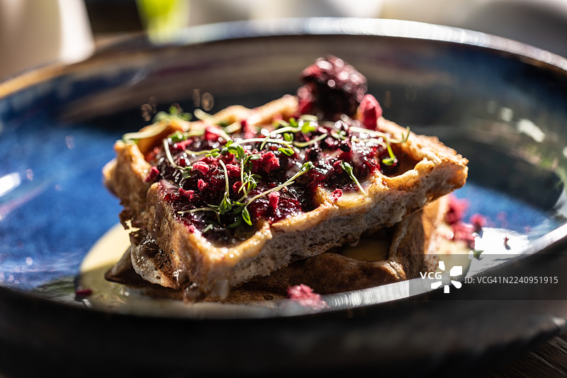 亮蓝色的闪亮碗里盛着华夫饼，华夫饼的两层上面淋着浅奶油或酱汁，并用深红色的撒料（可能是覆盆子或黑莓）和新鲜香草或嫩芽精心装饰。图片素材