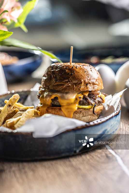 一份丰盛的牛肉汉堡，配融化的奶酪，用乡村风格的蓝白瓷碗盛装，旁边配有炸薯条。汉堡配有撒满芝麻的面包，用木串固定，周围环绕着酥脆的炸薯条，炸薯条放在一张白纸上。图片素材