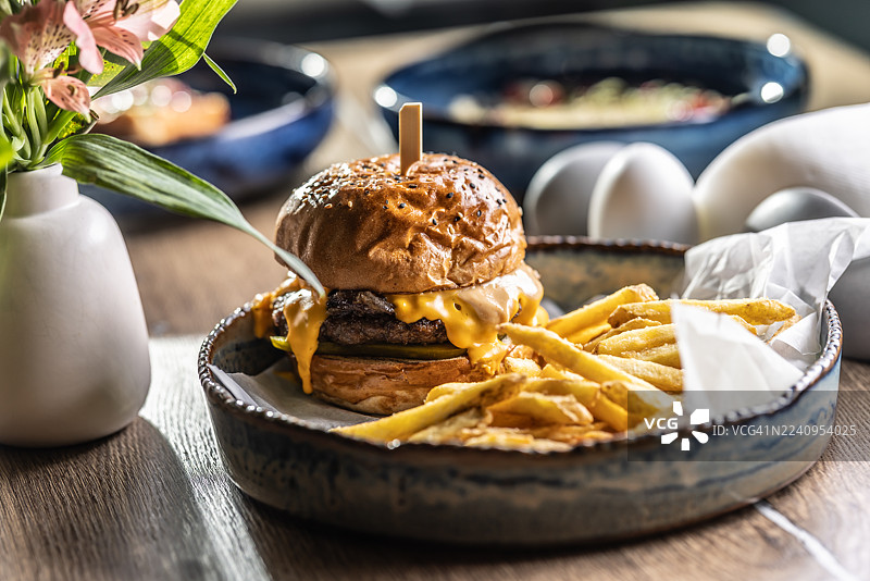 一份丰盛的牛肉汉堡配融化的奶酪，搭配盛放在乡村风格蓝白陶瓷碗里的炸薯条。汉堡配有撒有芝麻的面包，用木签串着，周围环绕着酥脆的炸薯条，薯条放在一张白纸上。图片素材