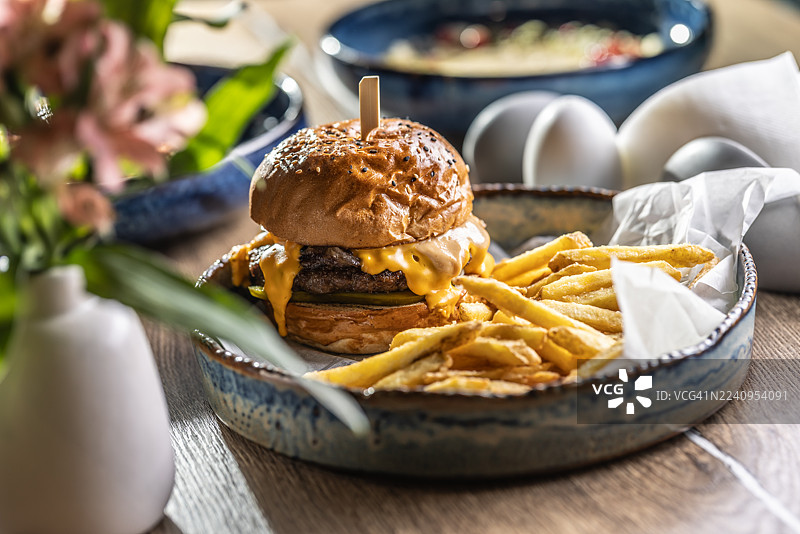 一份丰盛的牛肉汉堡，配有融化的奶酪，盛放在乡村风格的蓝白色陶瓷碗中，旁边是炸薯条。汉堡配有撒有芝麻的面包，用木串固定，周围环绕着酥脆的炸薯条，薯条放在一张白纸上。图片素材