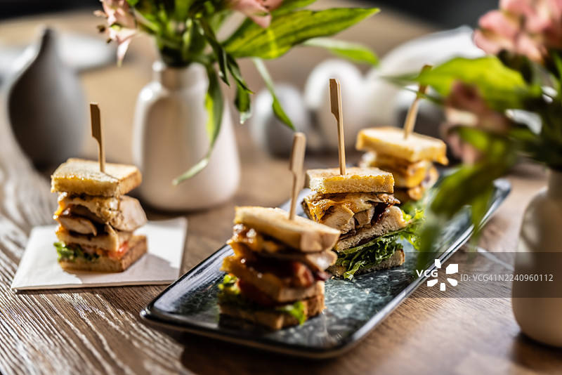 一只摆放在深色长方形亮面盘子里的三层俱乐部三明治，里面有鸡肉、培根和蔬菜，被切成四块，并用木签串好。背景中还有其他三明治和餐桌装饰，均处于失焦状态。图片素材