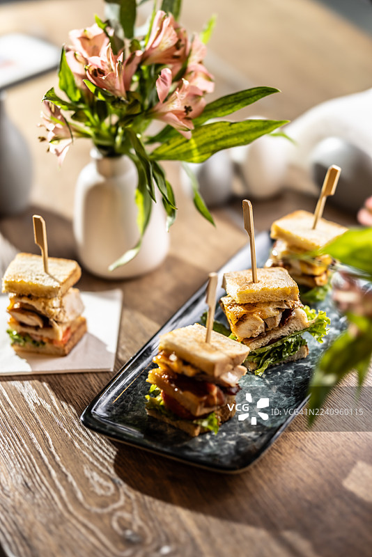 一只装有鸡肉、培根和蔬菜的三层俱乐部三明治，切成四块，用木串固定。三明治放在一个光亮、长方形的深色盘子上，背景中模糊地可以看到其他三明治和餐桌装饰。图片素材