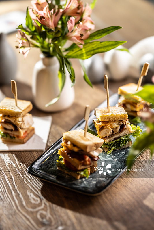 一只三层俱乐部三明治，包含鸡肉、培根和蔬菜，被切成四块，并用木串固定。三明治放在一个光亮的、矩形的、深色盘子上，背景中是模糊的其他三明治和餐桌装饰。图片素材