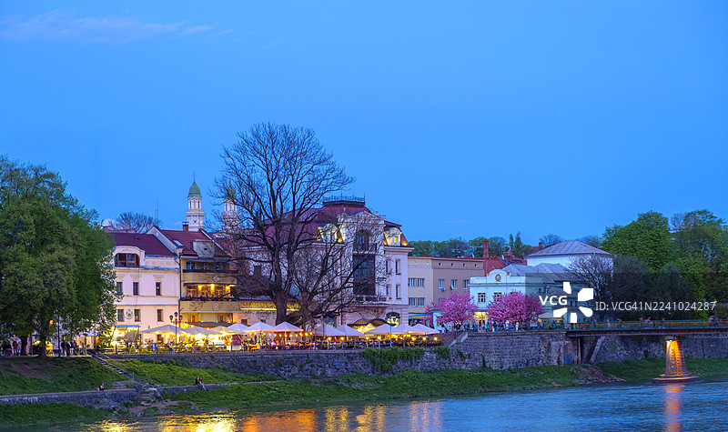 暮色中乌日霍罗德的城市灯光倒映在乌日河上，滨河餐厅和悠闲散步的人们在盛开的樱花树下享受着夜晚的美好时光图片素材