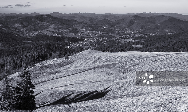 从喀尔巴阡西部乌克兰地区，一个布满纹理的草坡上远眺，山谷中座落着村庄，周围环绕着连绵的山峦和茂密的常绿森林。图片素材