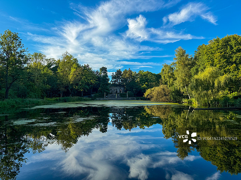 宁静的湖面倒映着蔚蓝的天空和缕缕白云，阳光明媚的一天，四周环绕着郁郁葱葱的绿树。图片素材
