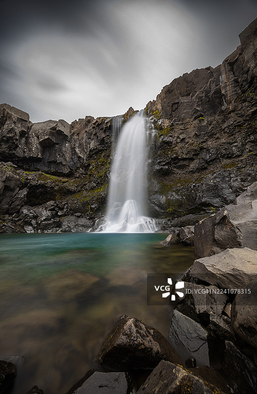 冰岛，在阴沉的天空下，雄伟的长曝光照片展示了强大的瀑布倾泻入宁静的水池，周围环绕着崎岖的黑色岩石。图片素材