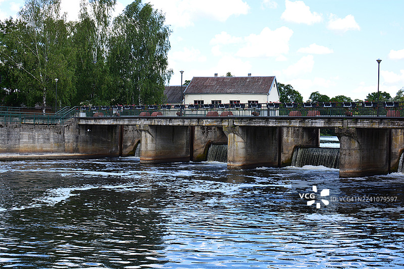 立陶宛考纳斯，风景优美的混凝土桥，桥下流水潺潺，四周绿树成荫，远处有一栋房屋，天空布满云彩。图片素材