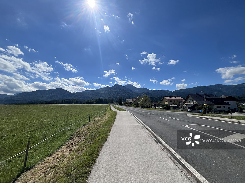 晴朗的一天，一条乡村公路经过房屋，后面是群山和田野，天空湛蓝，地点是奥地利菲拉赫（Villach）。图片素材