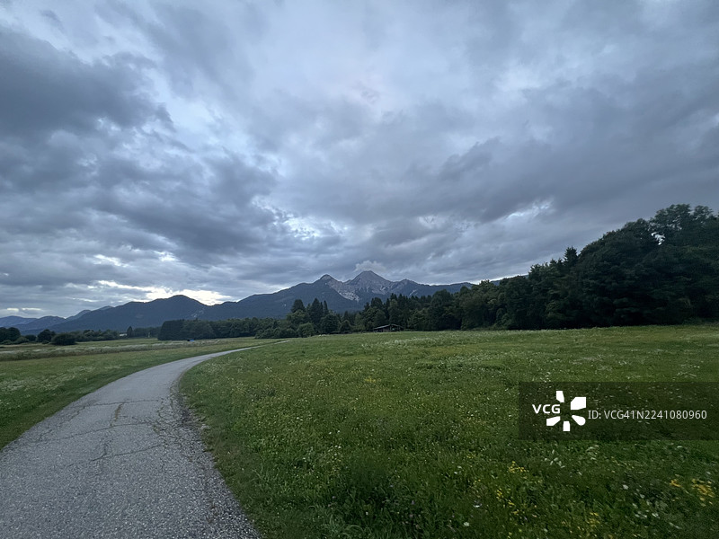 奥地利菲拉赫，一条蜿蜒的道路穿过绿色的田野，背景是山脉和戏剧性的多云天空图片素材