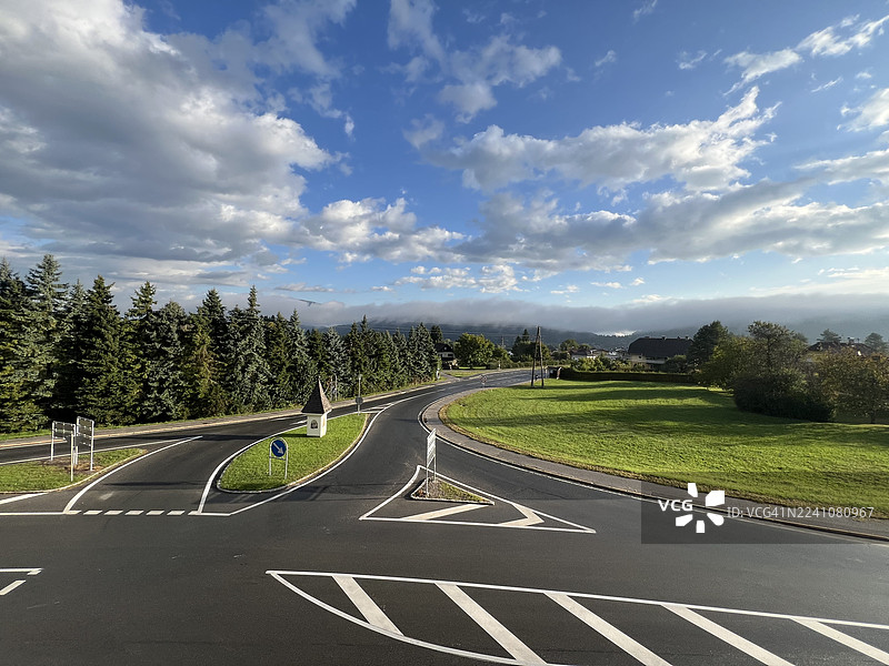 乡间空旷的道路和交叉口，有绿色的田野和森林，头顶是多云的蓝天，地点是奥地利菲拉赫。图片素材