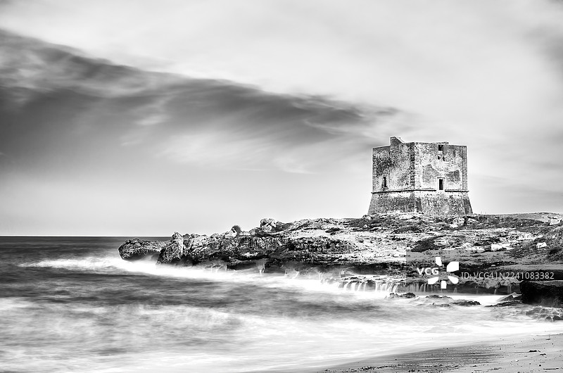 黑白照片：古老的瞭望塔矗立在岩石海岸上，下方海浪拍打，天空布满戏剧性的云彩。图片素材