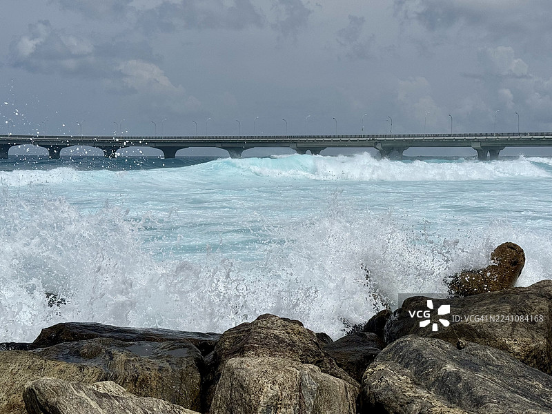 背景桥梁下，戏剧性的海浪拍打着岩石，天空布满乌云。图片素材