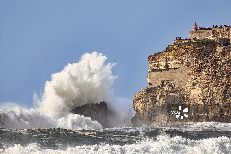 巨大的海浪拍打着悬崖，悬崖顶端有古老的堡垒和灯塔，天空湛蓝，葡萄牙图片素材
