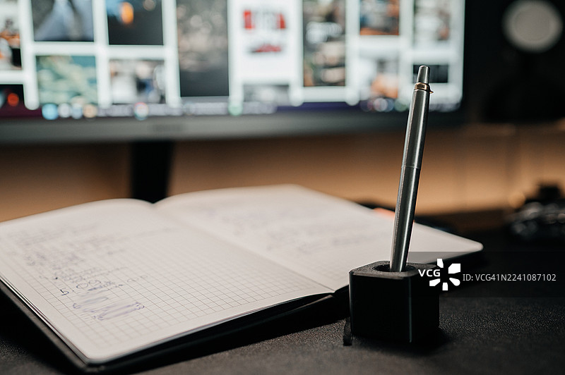 靠近打开的笔记本，桌上的笔筒里放着一支笔，背景是模糊的电脑显示器，暗示着工作或学习。图片素材
