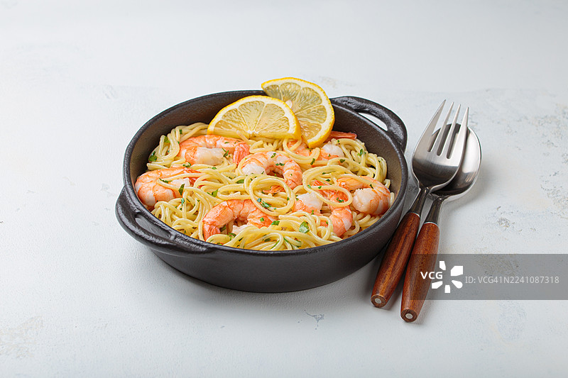 黑碗里盛着美味的鲜虾意面，配有新鲜柠檬片和欧芹，旁边放着餐具，背景为浅色。图片素材
