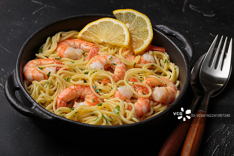 高角度拍摄的美味鲜虾意面，配有柠檬，盛放在铸铁锅中，放在深色桌子上，准备上菜。图片素材