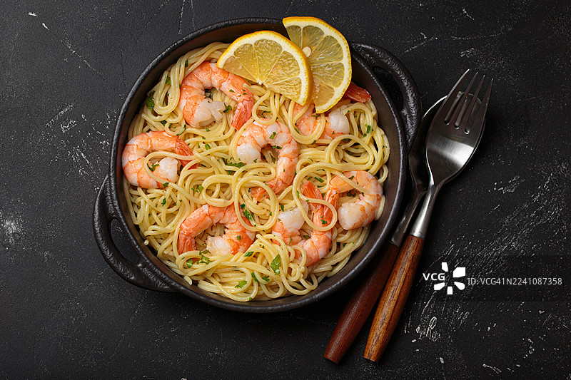 美味的鲜虾意大利面配柠檬片和新鲜欧芹，盛放在黑碗中，配有餐具，置于深色背景上图片素材