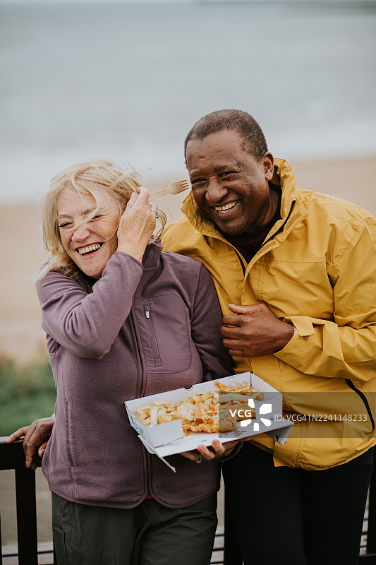一对成熟的夫妇在沙滩上分享一份炸鱼薯条，海风吹乱了女人的头发，他们看着镜头大笑。图片素材