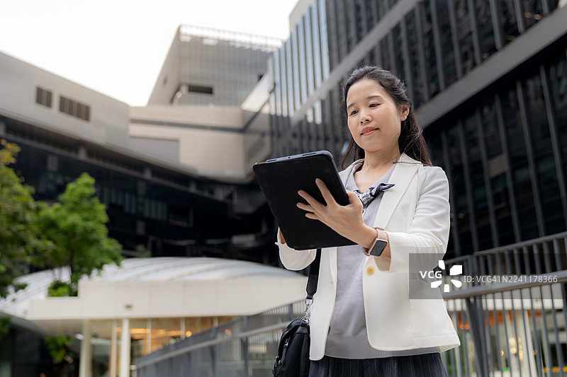 一位自信的亚洲企业家，手持平板电脑站在户外，在城市建筑的环绕下记录新想法。象征着现代企业生活方式和幸福感中的雄心、创新、创业精神和成功。图片素材