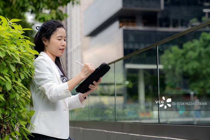 现代亚裔经理在绿色办公楼附近，在平板电脑上查看商业数据并记录想法。代表商业金融、电子商务、ESG创新、数字经济和科技驱动工作中的可持续领导力。图片素材