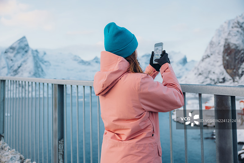 身穿粉色夹克的旅行者正在拍摄罗弗敦群岛上，红色的渔村和白雪皑皑的山脉组成的壮丽冬季景观图片素材