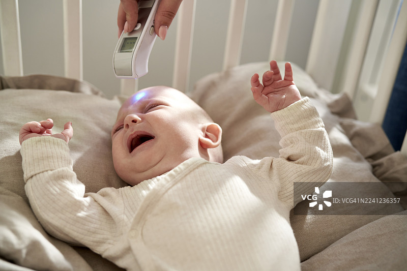 妈妈在家坐在婴儿床边，检查哭闹宝宝的体温。图片素材