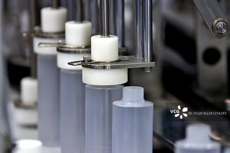 自动化灌装和封盖过程。精密机械夹持透明塑料瓶进行封盖或密封，强调化妆品或制药厂无菌高效的生产流程。图片素材