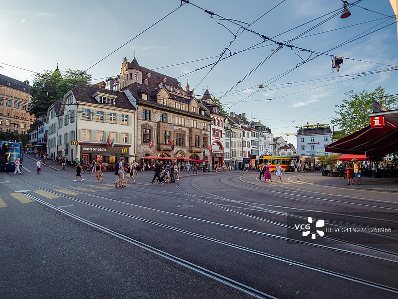 傍晚时分，瑞士巴塞尔城市中心的巴尔富瑟广场（Barfüsserplatz）的广角景观，可以看到电车轨道和电缆，行人或在咖啡馆露台上悠闲享受，以及美丽的古老建筑。图片素材