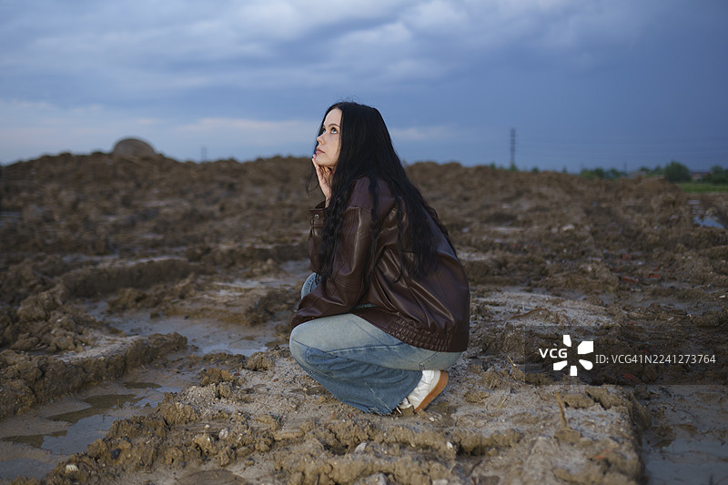 暮色中，身穿皮夹克的年轻女子坐在泥土田野上图片素材