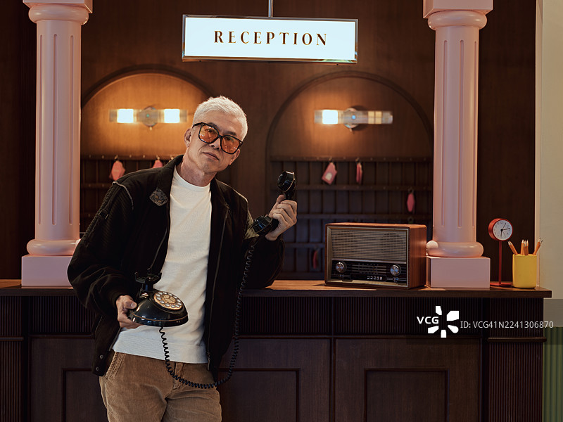 时尚的亚洲中年男性在复古酒店接待台处手持老式转盘电话。图片素材