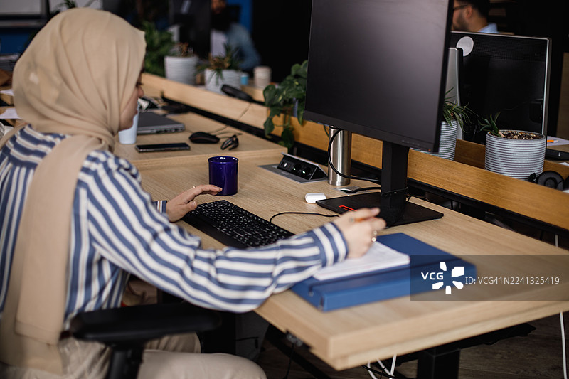 身着头巾的女性在现代办公室的办公桌前工作，桌上有显示器、笔记本和植物图片素材