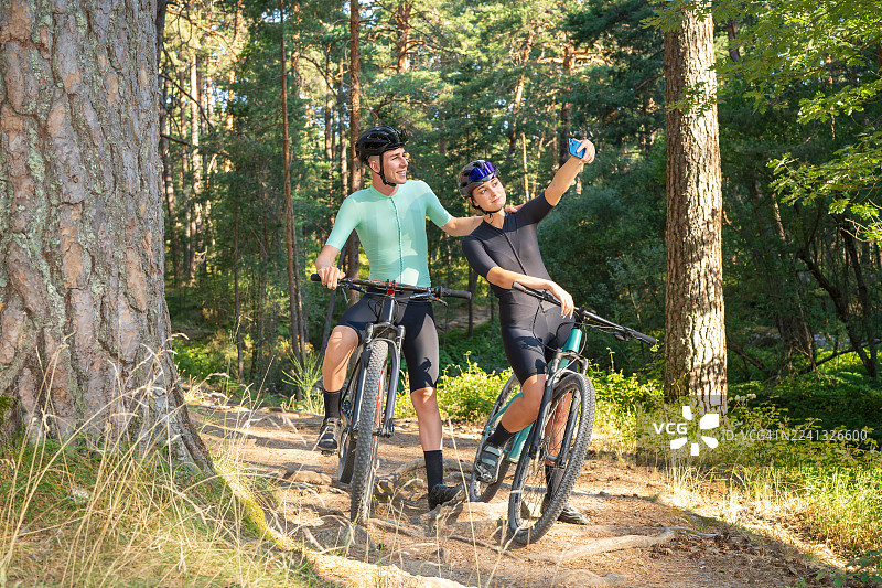 两位年轻的山地车手,一男一女,在骑行后于森林小径上停下来,兴高采烈地自拍,享受户外探险、健身和彼此的陪伴。地点:西班牙卡斯蒂利亚-莱昂塞戈维亚的皇家圣伊尔德丰索庄园。图片素材
