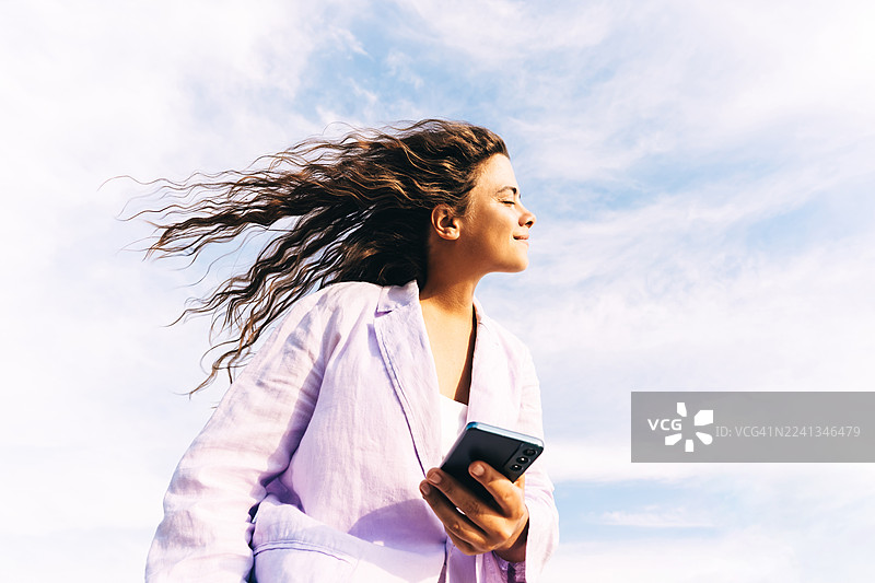一位年轻女子身穿浅紫丁香色夹克，在明媚的天空下看着智能手机，脸上带着微笑。图片素材