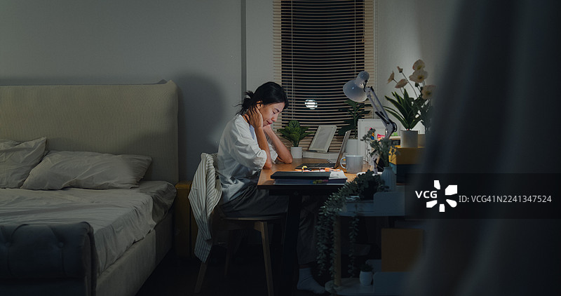 年轻的亚洲女性自由职业者晚上在家卧室用笔记本电脑工作到很晚，显得压力重重、筋疲力尽。远程工作和居家办公概念。图片素材