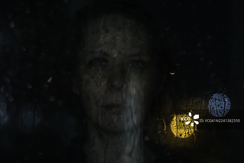 夜色中，一位忧思的女性透过布满雨痕的窗户向外凝望。图片素材