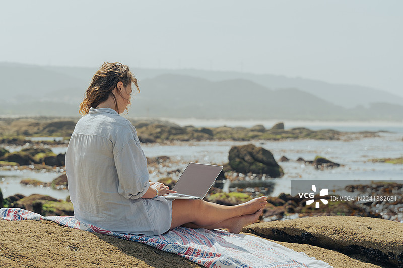 年轻女子在海边岩石上用笔记本电脑远程工作——海滩上的数字游民生活方式图片素材